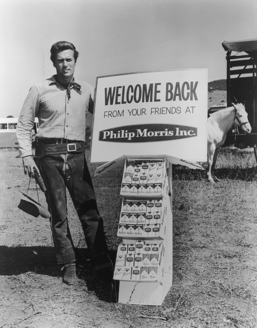 circa 1965: Full-length portrait of American actor Clint Eastwood posing in Western gear next to a display of Philip Morris tobacco products in front of a white horse on an outdoor ranch. Eastwood starred in the television series, 'Rawhide'. He wears chaps, a gun holster, and a bandana while a cowboy hat hangs by his side. (Photo by Hulton Archive/Getty Images)