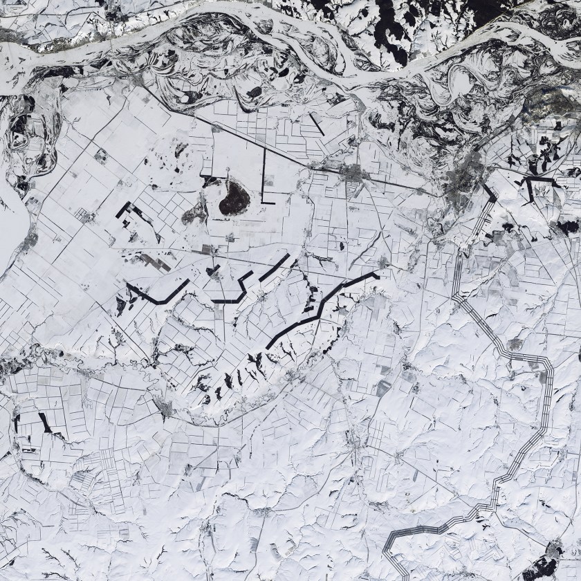 Is this a black-and-white image? No, this is a natural color image of snow-covered southwestern Russia. Windbreaks, roads, and fence lines look like random pencil marks near the Volga River, which flows across the top of the image. The thick lines are trees planted to protect fields from dry wind and erosion; these windbreaks retain snow, allowing more moisture to penetrate into the soil. (USGS/NASA)