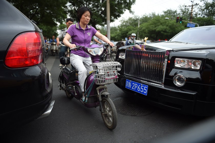 A woman squeezing her electric scooter between a Rolls-Royce (R) and Porsche SUV at a busy intersection in Beijing. The new tax took effect December 1, 2016 and was intended to "guide rational consumption" and promote energy-efficient vehicles, the finance ministry said in a statement late November 30, 2016. (GREG BAKER / AFP)