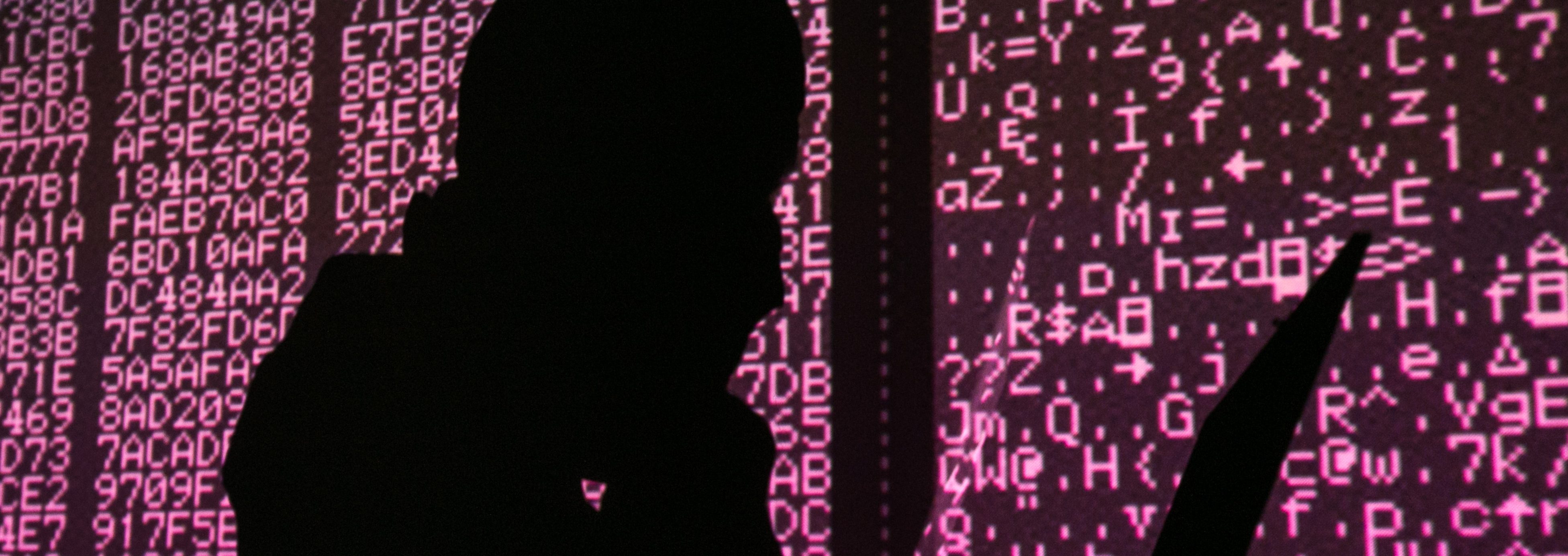 A silhouette of a man in a balaclava mask sitting at a laptop computer, with computer code in the background. Sergei Konkov/TASS (Photo by Sergei KonkovTASS via Getty Images)