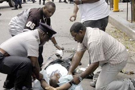 Police and pedestrians help a man wounded 11 July, 2005 after an explosive artifact exploded in a busy shopping district in downtown Port-of-Spain, Trinidad and Tobago, wounding 13 people. Police have yet to confirm if the blast was caused by a bomb or a hand grenade thrown into the bin, as some eyewitness reported.  AFP PHOTO STR   MAXIMUM QUALITY AVAILABLE  TRINIDAD AND TOBAGO OUT  (Photo credit should read STR/AFP/Getty Images)