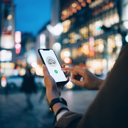 Young woman using mobile app on smart phone to arrange taxi ride in downtown city street, with illuminated city traffic scene as background