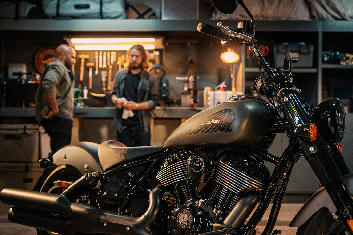 A bike from Indian Motorcycles with the Native American headdress logo on the gas tank sits in the foreground with two men in the background