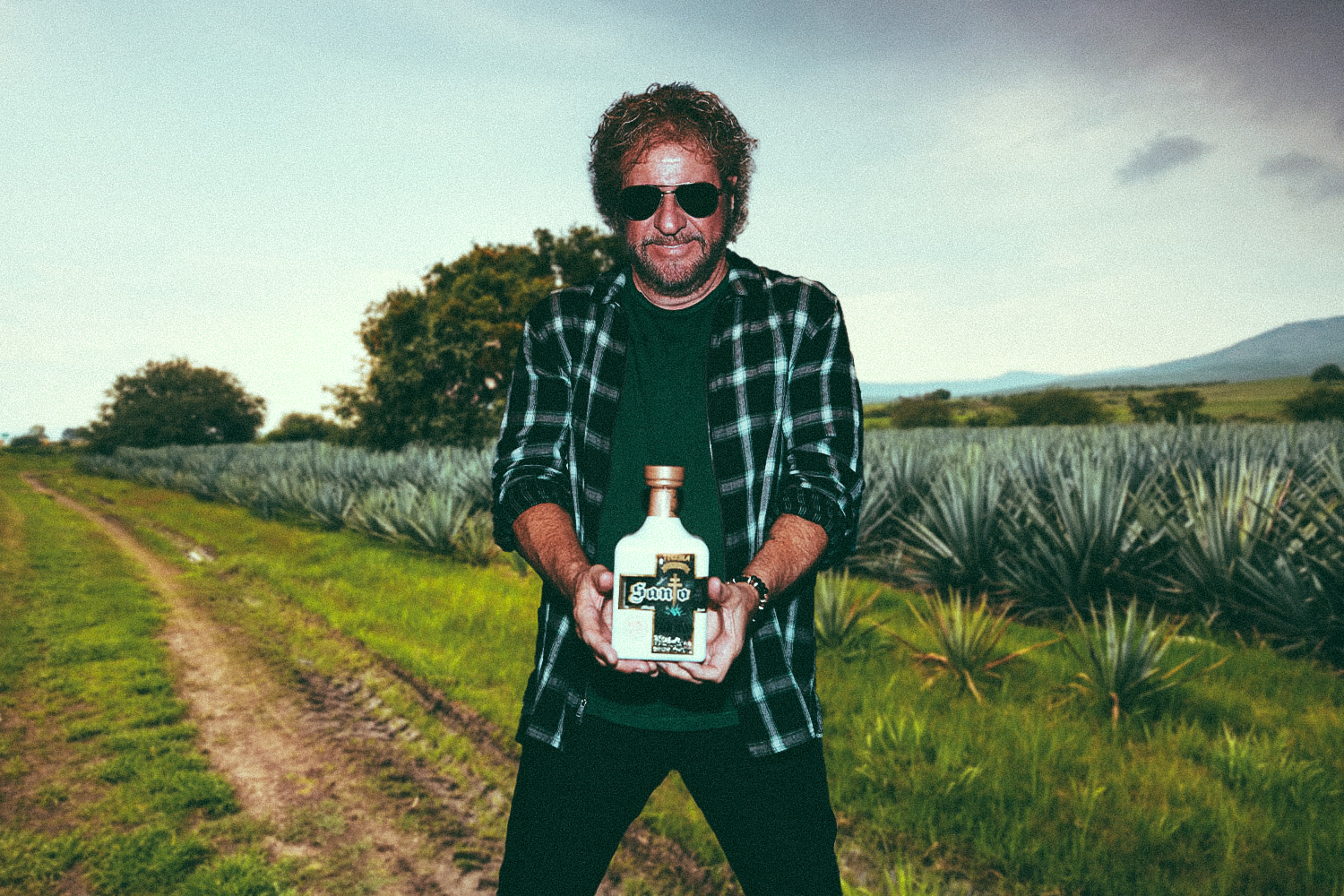 A composite image of Sammy Hagar with a bottle of this Santo tequila, superimposed on an agave field