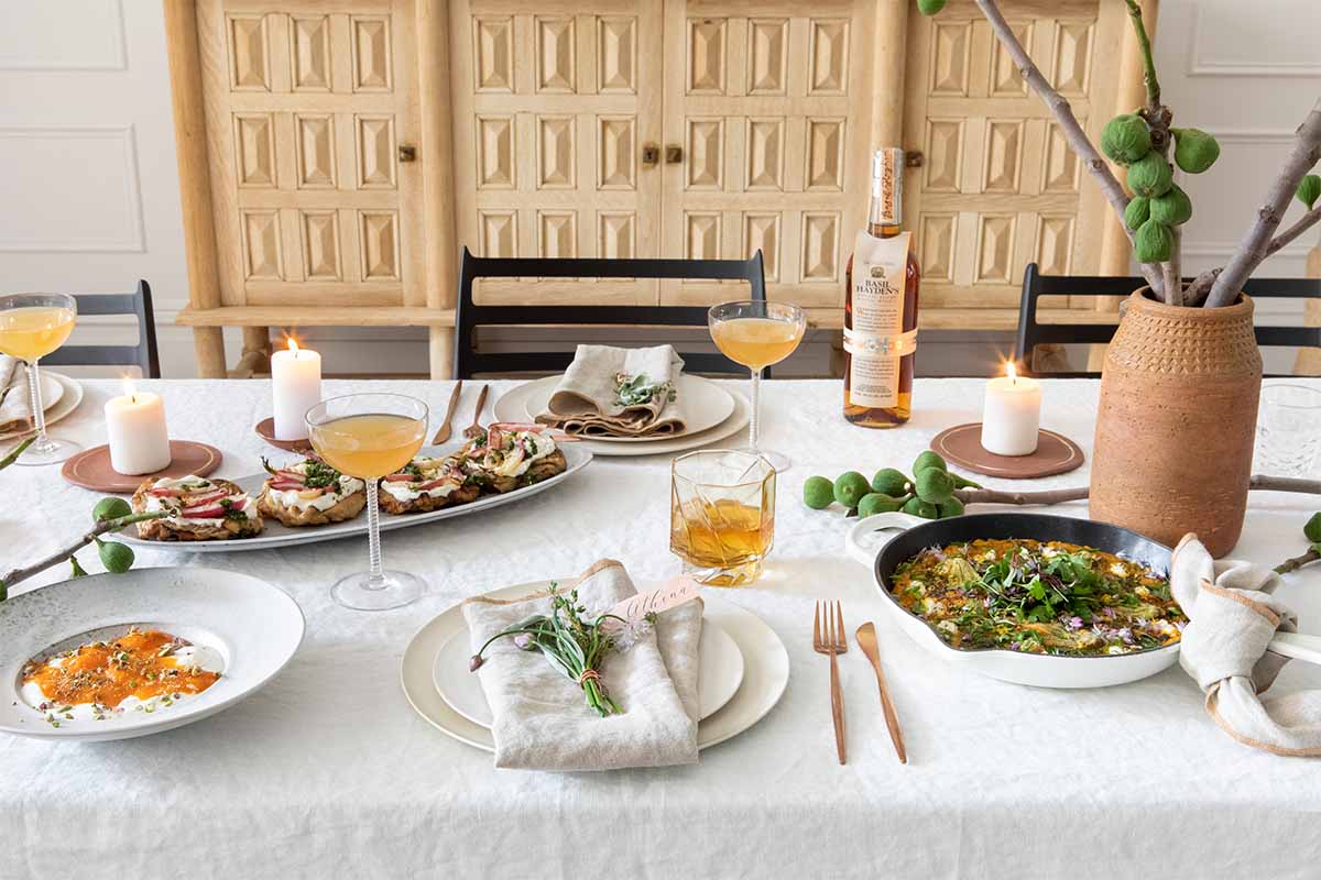 A table with brunch food, place settings, multiple cocktails and a bottle of Basil Hayden bourbon. Unconventional brunch drinks can improve your easy morning dining experience.