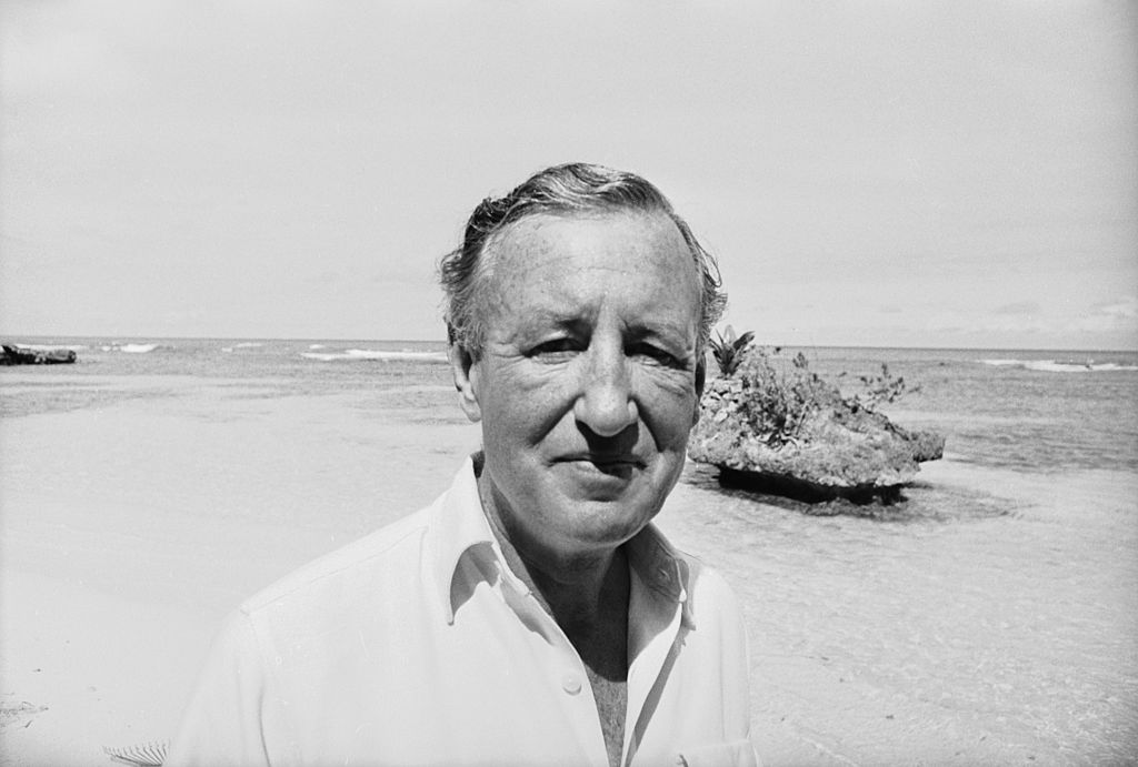 A black and white photo of author Ian Fleming on a beach. We take a look at how his wartime espionage work influenced writing his James Bond books.