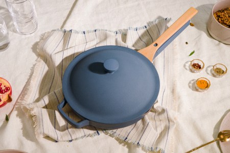 A blue Always Pan from cookware brand Our Place sitting in the middle of a table surrounded by glasses, spices, plates and a pomegranate