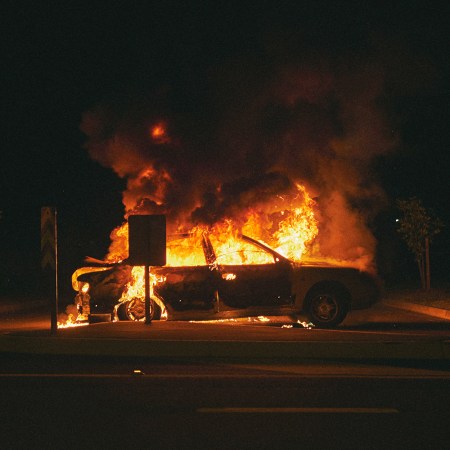 A four-door car on fire in a parking lot at night, with black smoke billowing up into the night sky