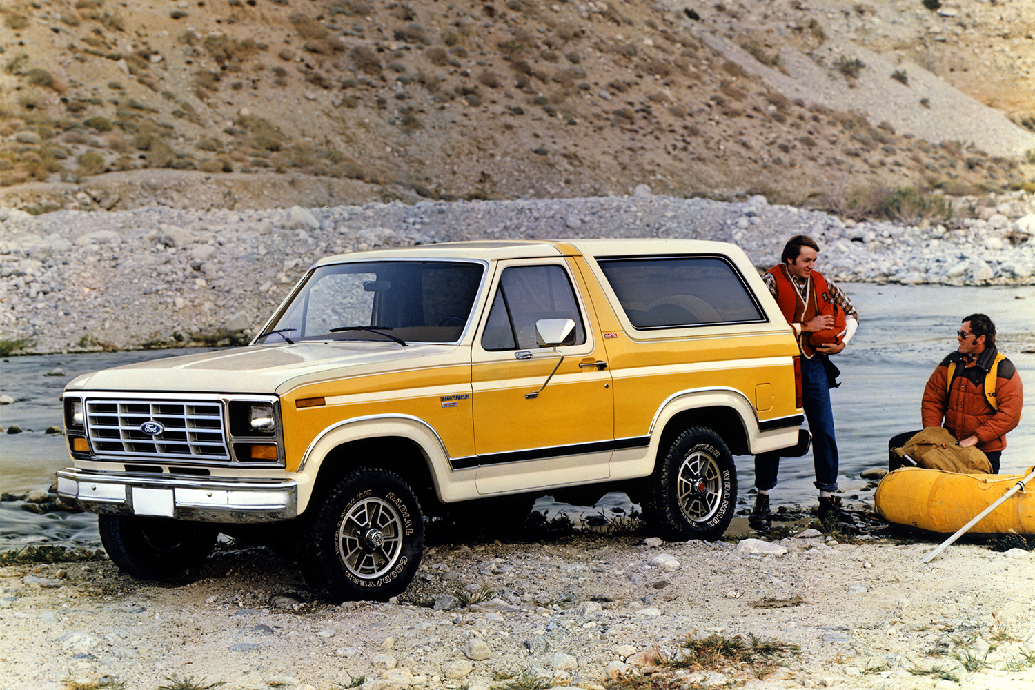 A white and yellow 1982 Ford Bronco SUV sitting among rocks and a river next to two outdoorsmen in a retro photo