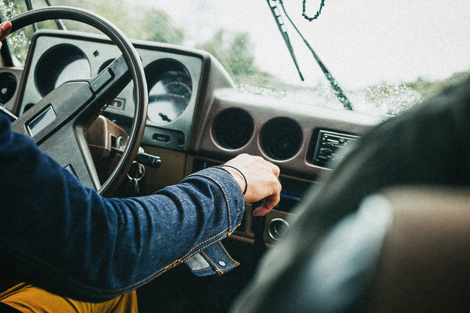 A man in a jean jacket in a car with his hand grasping the stick shift.