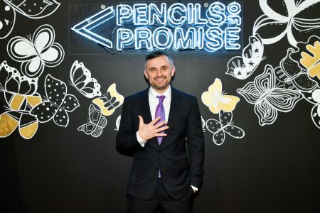 Entrepreneur Gary Vaynerchuk in a black suit with a white shirt and purple tie standing in front of a neon sign that says "Pencils of Promise"