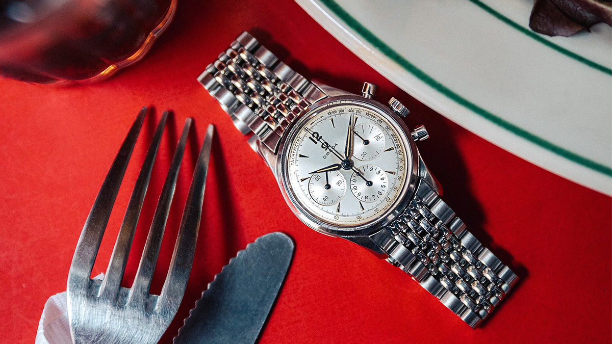 A vintage Omega Seamaster Chronograph sitting on a red tablecloth on a dining table next to a fork, knife and plate. The vintage watch is currently on sale through J.Crew.