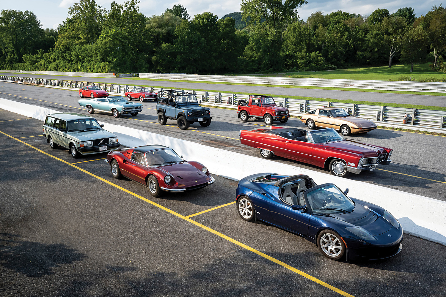 A race track with collector cars lined up, including the Tesla Roadster Sport, Ferrari 246 Dino, Land Rover Defender and others. They're all on the Hagerty Bull Market List for 2022.