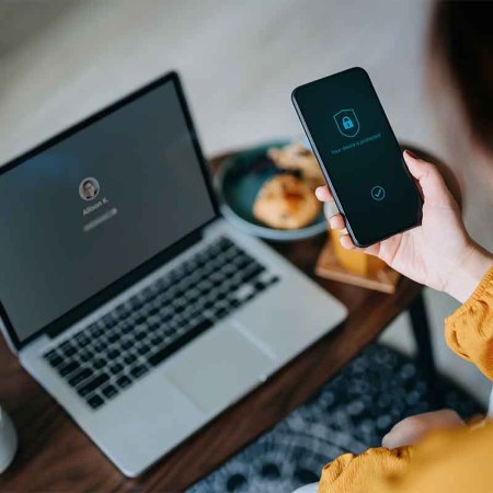 stock photo of Young Asian woman logging in to her laptop and holding smartphone on hand with a security key lock icon on the screen, sitting in the living room at cozy home. Password security for corporations was recently called out by Dashlane.