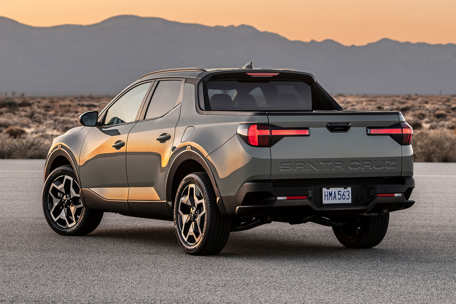 The 2022 Hyundai Santa Cruz pickup truck in grey sitting at a standstill. This photo shows the rear 3/4 of the pickup with the lights on.