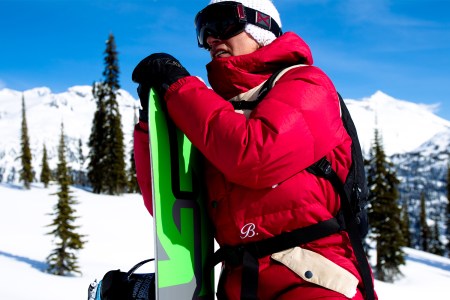 Jake Burton, the legendary snowboarder, standing on a snowy mountain holding a snowboard with goggles and a hat on. We spoke with Jake's friends and colleagues about the new documentary Dear Rider ahead of "A Day for Jake."