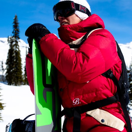 Jake Burton, the legendary snowboarder, standing on a snowy mountain holding a snowboard with goggles and a hat on. We spoke with Jake's friends and colleagues about the new documentary Dear Rider ahead of "A Day for Jake."