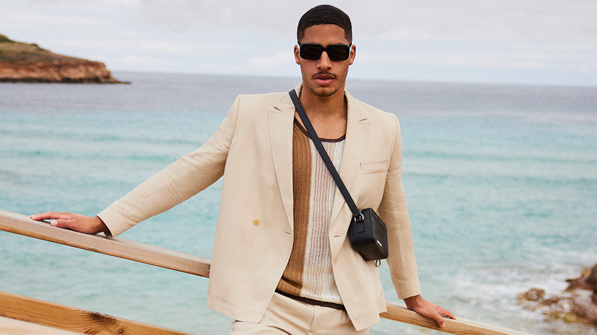 a model in a white suit, shirt and black leather bag standing in front of the ocean, courtesy of Mytheresa