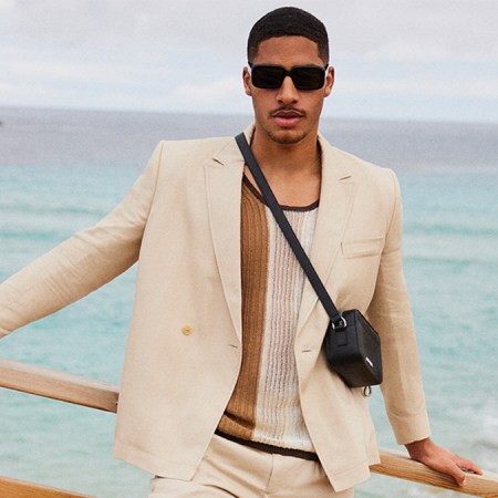 a model in a white suit, shirt and black leather bag standing in front of the ocean, courtesy of Mytheresa