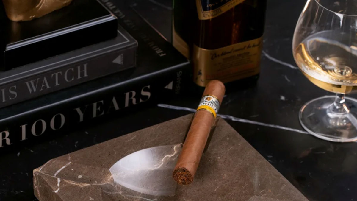 a stock shot of a cigar and ashtray, as well as books and whiskey, on a marble countertop