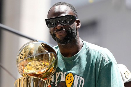 A photo of Golden State Warriors Forward Draymond Green wearing sunglasses and a green shirt and holding the 2022 NBA championship trophy