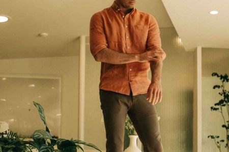 a model in the taylor stitch Jack linen long sleeve shirt and brown pants standing and rolling up his sleeves in a minimalist beige living room with lots of plants