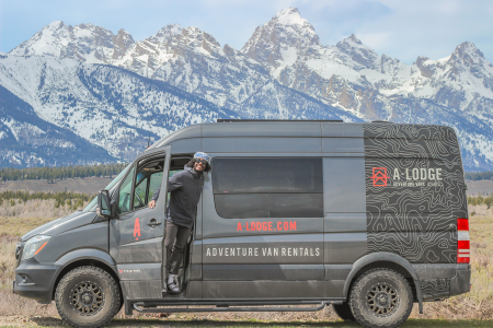 Writer and adventurer Joe Kanzangu stands in the door of his adventure van from A-Lodge with snow-capped mountain peaks in the background