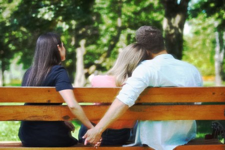 Three people sitting on a bench, one woman has her head resting on a man's shoulder, while the man hold hands with another woman.