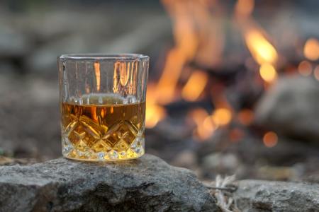 A glass of bourbon whiskey sits on a rock with a campfire in the background