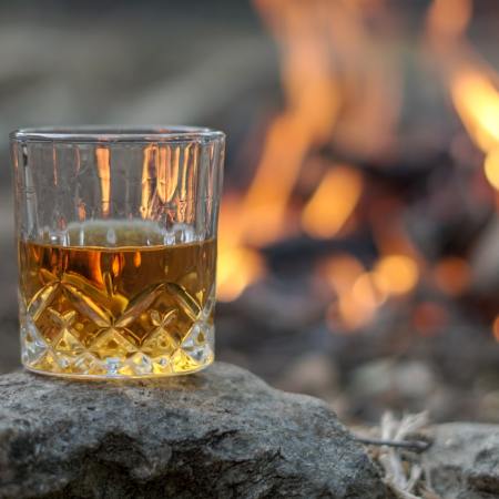 A glass of bourbon whiskey sits on a rock with a campfire in the background