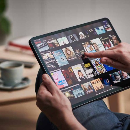 Detail of a mans hand scrolling through Netflix on an Apple iPad Pro. In certain countries, Netflix is cracking down on password sharing through a confusing new system.