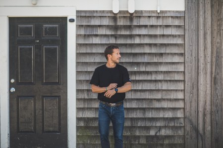 A man with a perfect tucked in black t-shirt standing in front of a grey wall.