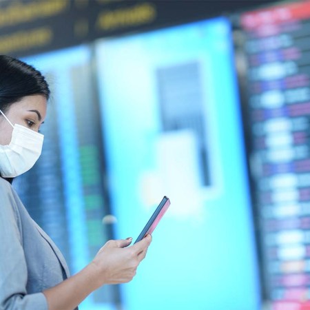 Woman wearing protective face mask checking flight schedule on mobile phone while standing in front of the arrival departure board. A new Twitter scam has fraudulent airline accounts attempting to take money from passengers