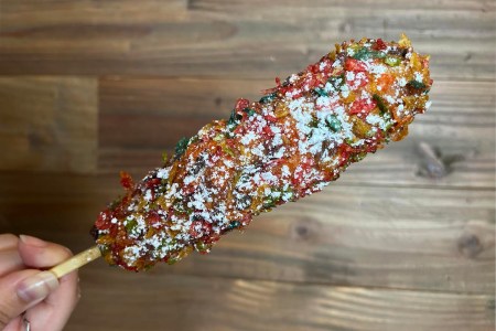 A person holding a Rainbow Kong Dog, a corn dog with fruity rice puffs and powdered sugar. It's part of the sweet hot dog trend.