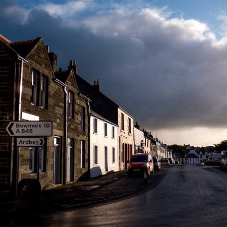 Looking down main street of Port Ellen sunlight with big storm clouds - stock photo. Peat supplies from Port Ellen may be curtailed in the next two years.