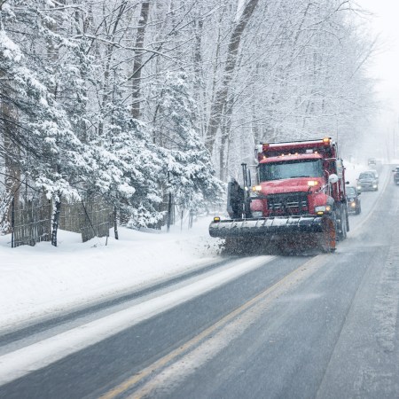 A red snowplow plowing snow up a hill on a steep rural road