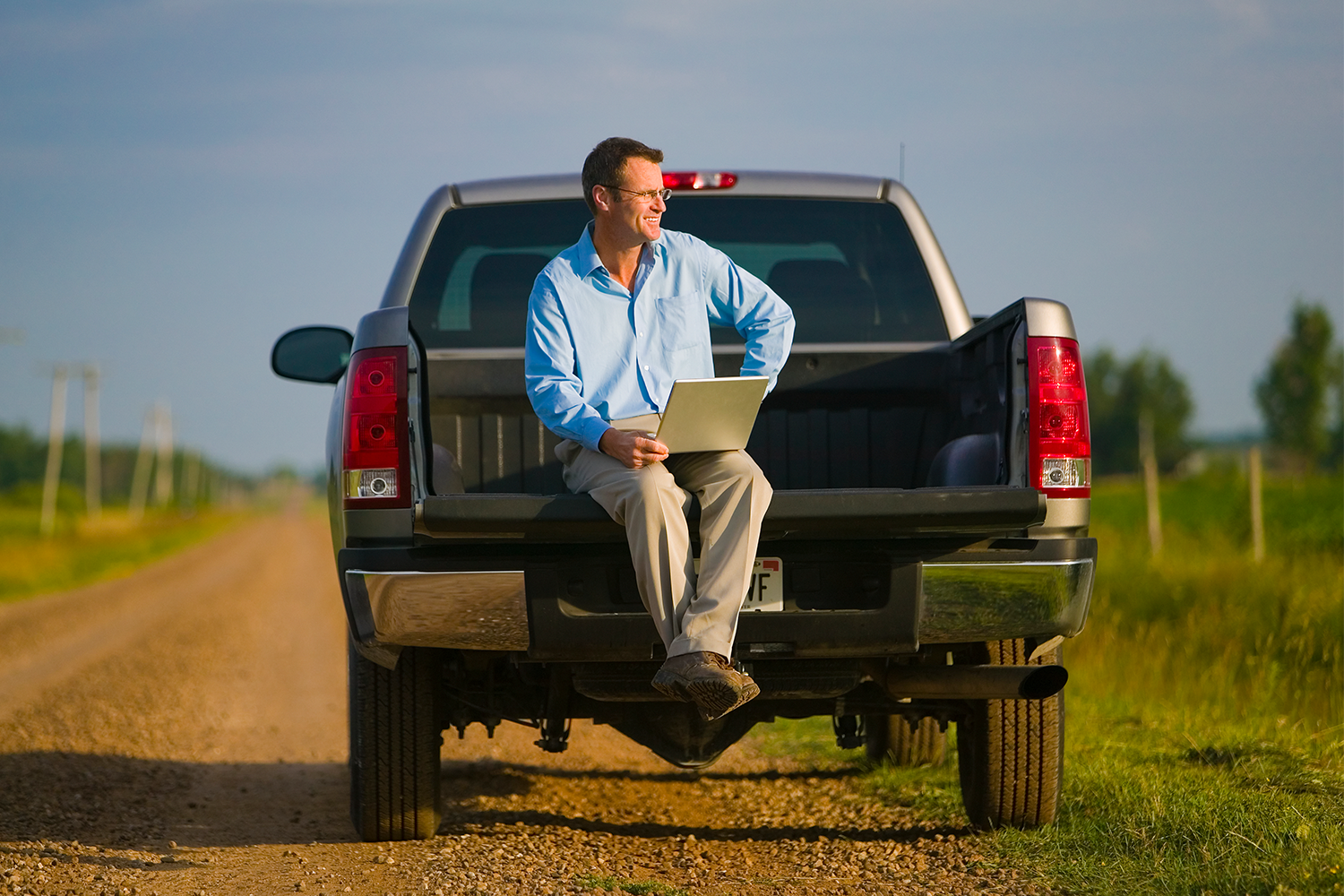 A man in a button-down shirt and khakis sitting in the bed of a pickup truck with a laptop computer. According to survey data, pickup truck buyers know they don't actually need their trucks.