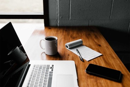 A laptop and coffee and a notepad on a table. Working out of office and/or at home is showing to be more productive for both workers and businesses.