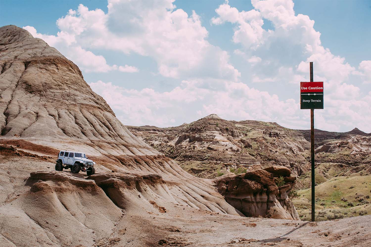 White Jeep Wrangler (remote control) off-road vehicle parked atop a cliff with a sign noting steep terrain and to seek caution.