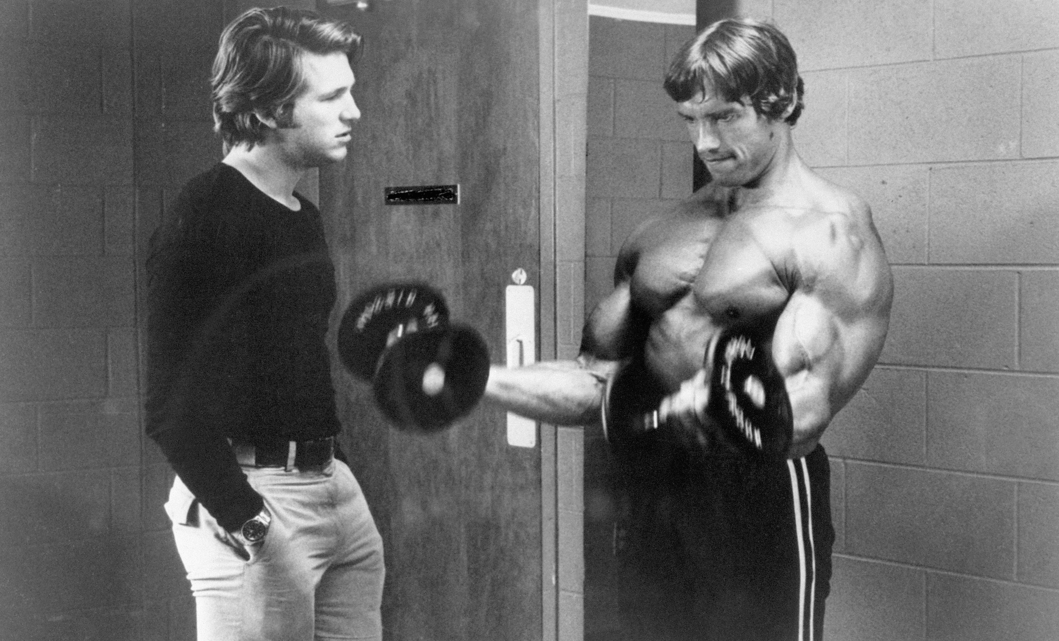 Jeff Bridges talking to a shirtless Arnold Schwarzenegger, who is curling a barbell.