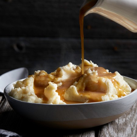 Jug of gravy being poured onto bowl of steaming mashed potatoes