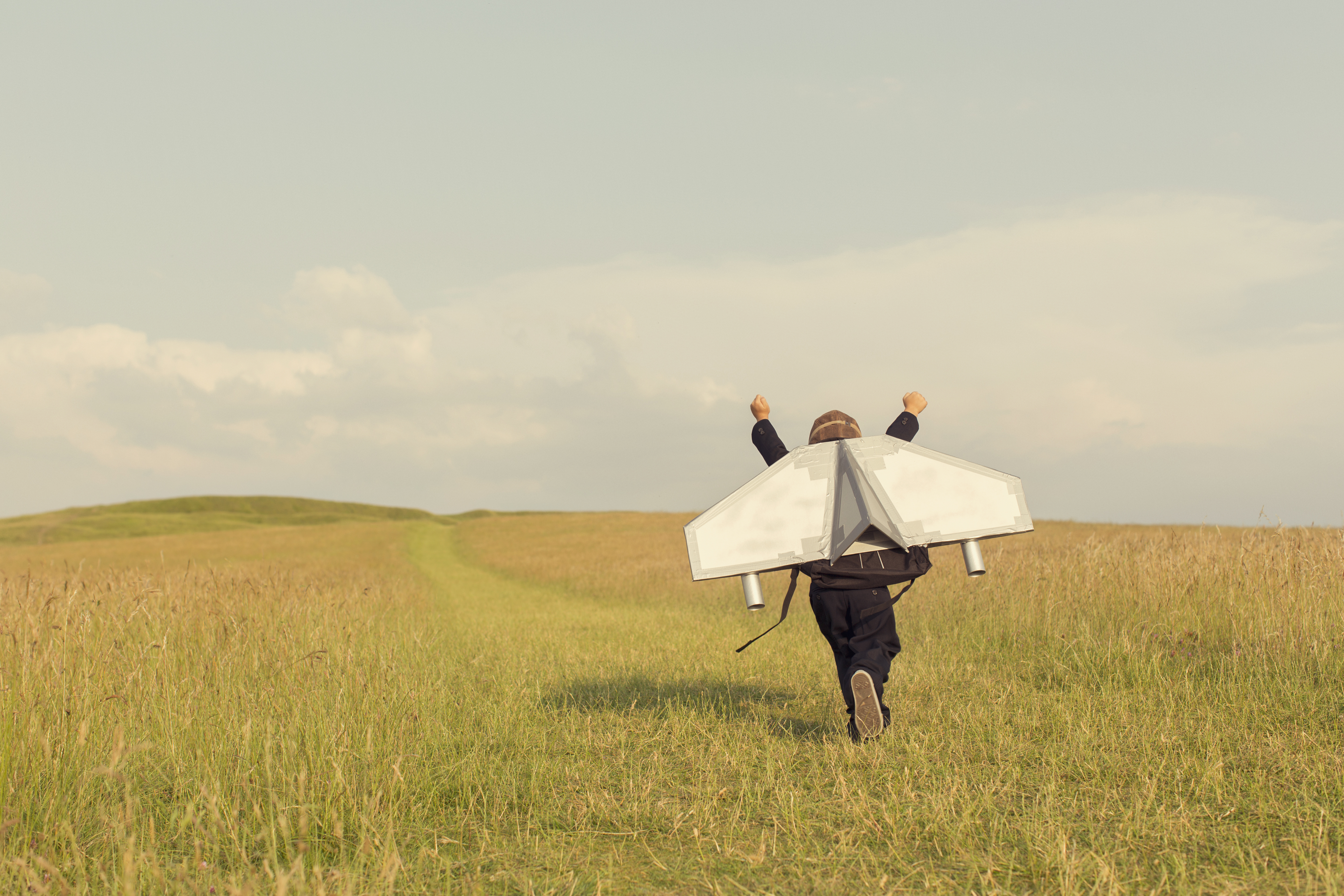 A kid running through a field with an arts and crafts jetpack on his back.